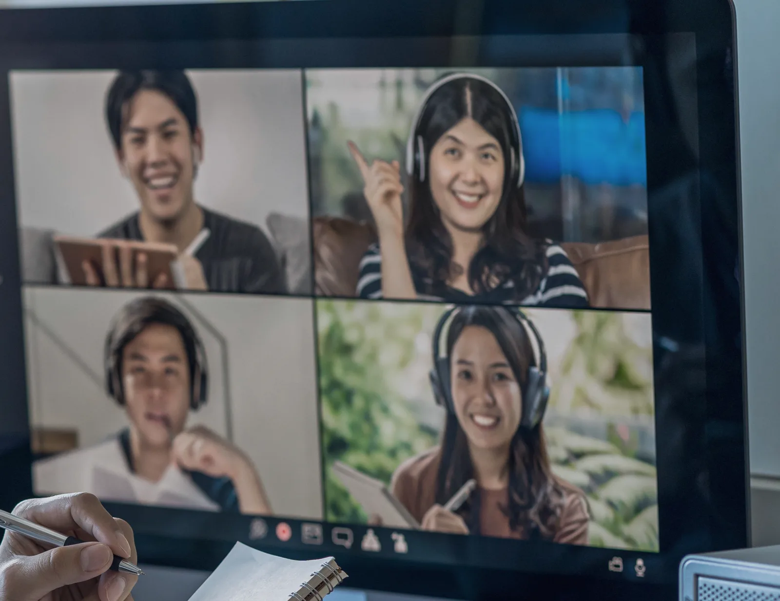 Video call screen showing four smiling remote colleagues, with a hand holding a pen in the foreground