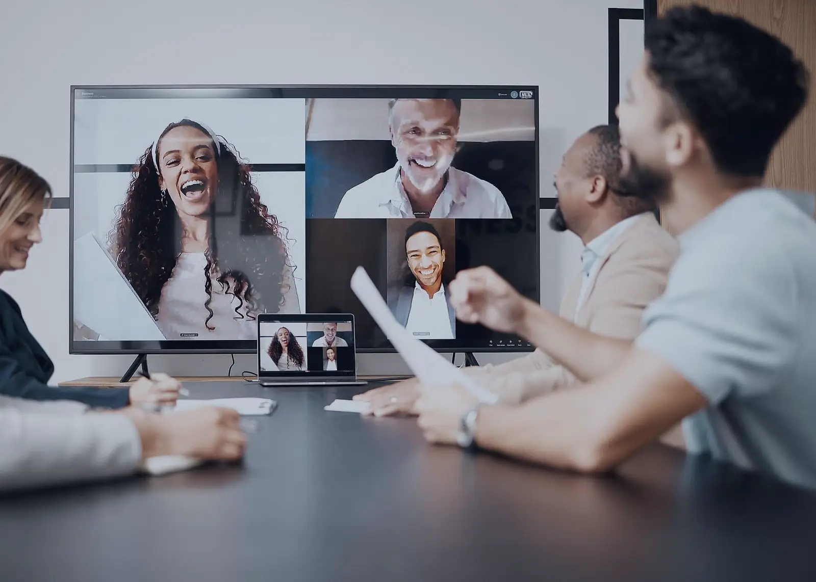 Colleagues in a meeting room joined by remote teammates on a video call screen showing four participants